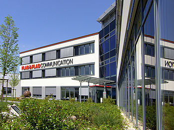 Gebäudefassade mit großen Fenstern und überdachtem Eingang. Davor Bepflanzung mit Bäumen, Wiese und Sträuchern.